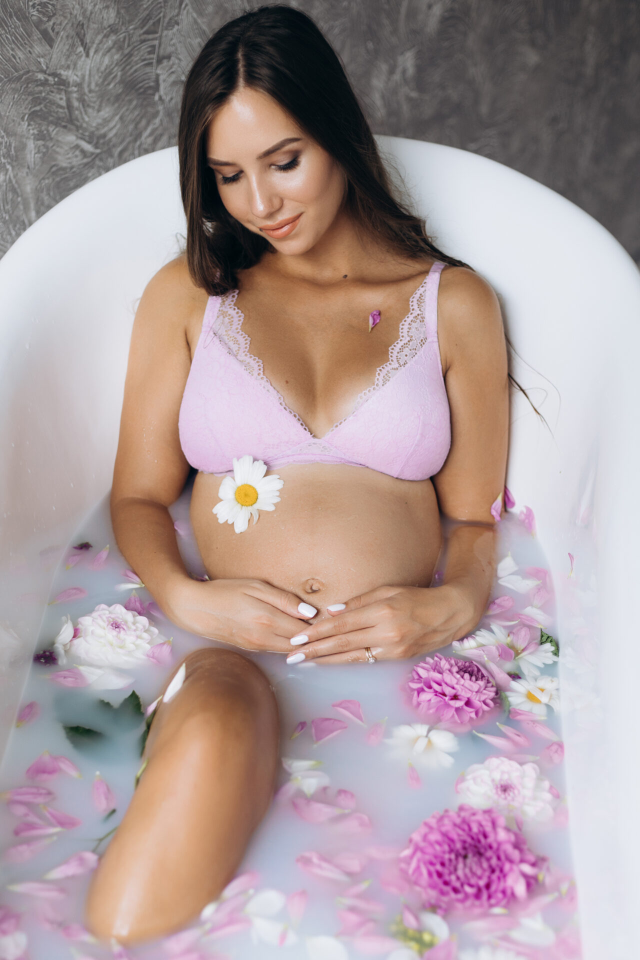 Sensetive portrait of an expecting woman. Beautiful pregnant woman in pink lingerie lies in a bath full of pink flowers and milk